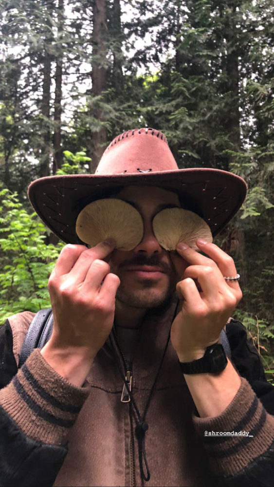 Alex Ross holds two mushroom caps up in front of his eyes. Text reads @shroomdaddy_