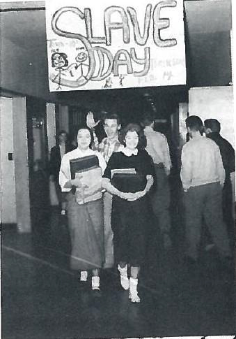 Historical photo of students walking in a hall at Chilliwack highschool in 1950s, with a large sign overhead that reads "Slave Day."