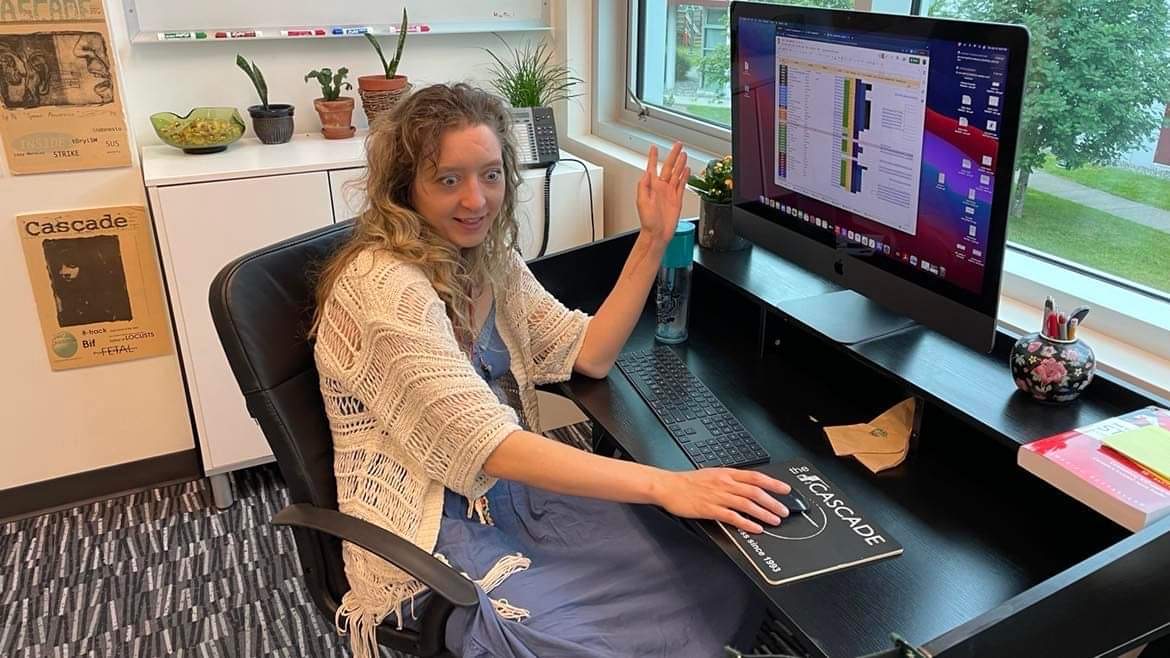 Photo of Andrea Sadowski sitting at a desk in The Cascade office, working at a computer and waving with a jokingly haunted look on her face
