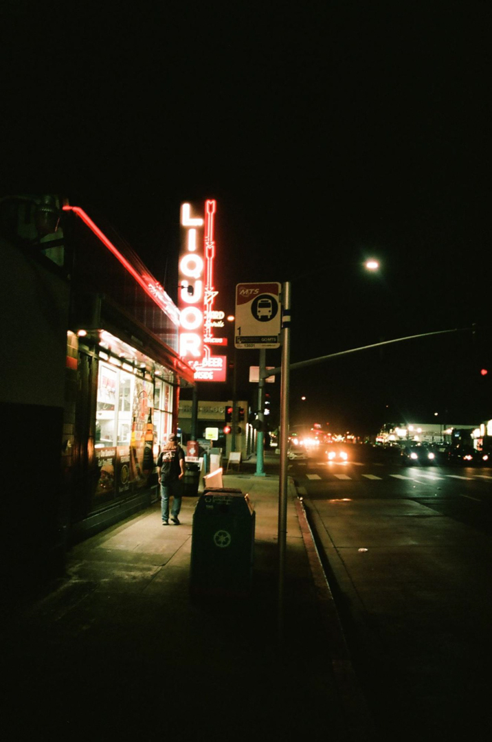 Photo of a dark street at night, light mainly by a neon liquor store sign, with a person walking the glow of the store's window.