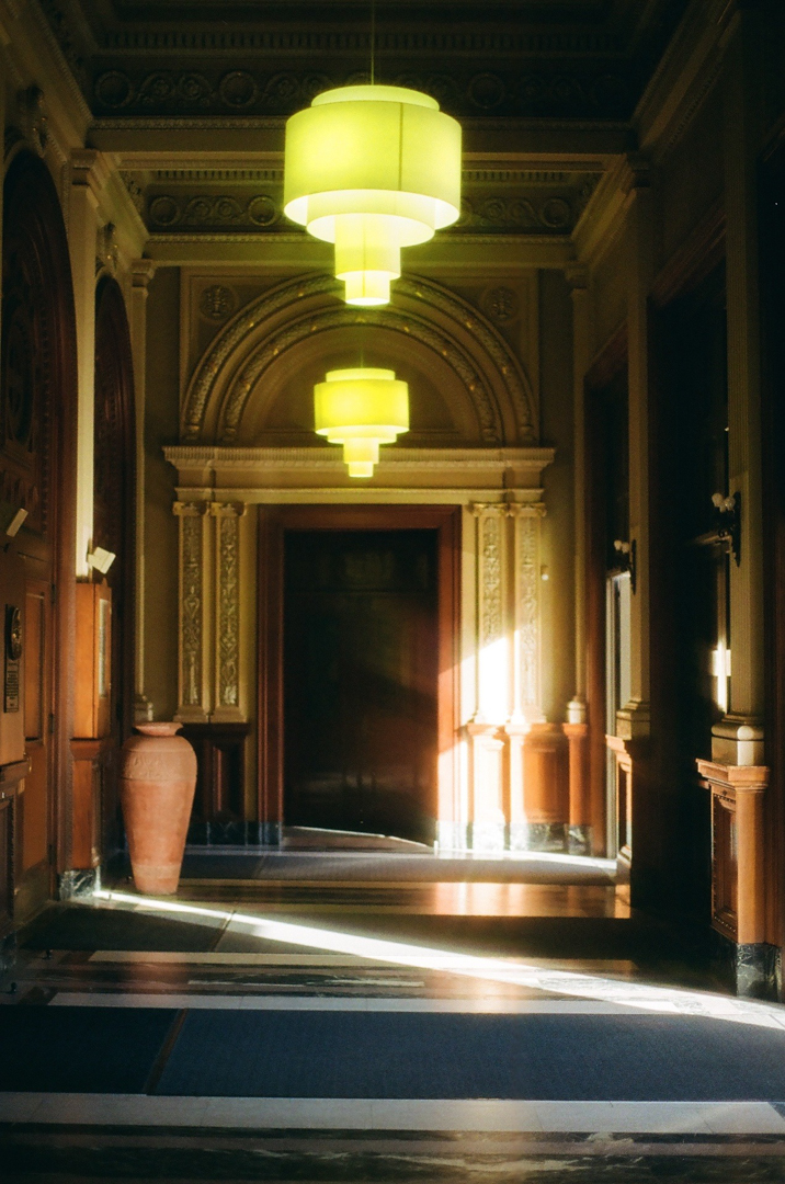 Photo of hallway in an old, ornate building. Hanging chandeliers are above, and a large old vase sits to one side. A dark doorway is centered at the opposite end of the hall.
