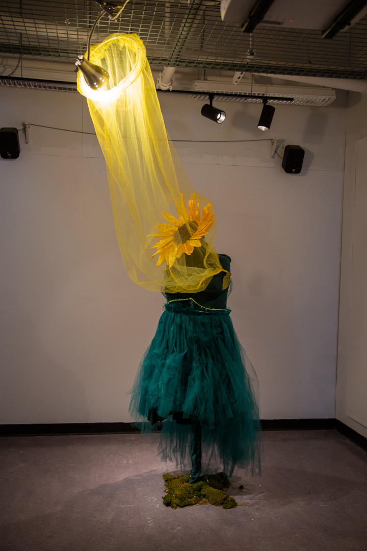 Photo of an art piece in a gallery. A green dress hangs on a mannequin, with a sunflower for a head. A light above illuminates it, and yellow fabric leads down from the light to the sunflower.