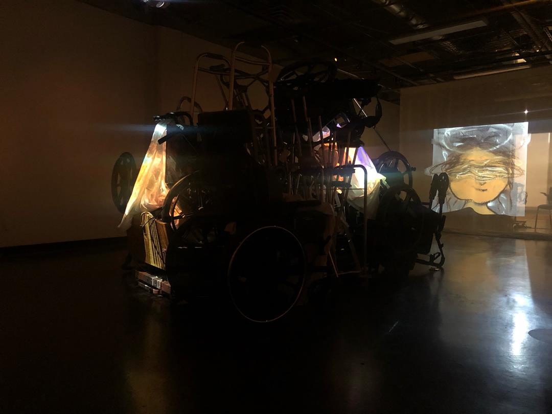 A dark art gallery room, in the center is a pile of equipment like a wheelchair and a walker. There is a projector in the center of the pile and it is projecting images onto the wall across. The images are paintings of abstract faces.