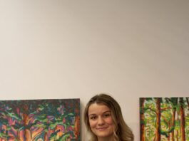 The artist Kiara Louwerse stading with her hadn on her hip in frommt of 2 of her artworks. The paintings are of trees in the forest.