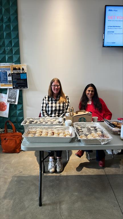 A photo taken at the event of the bakesale table with varous goods on it and sitting behind is two of the sales coordinators.