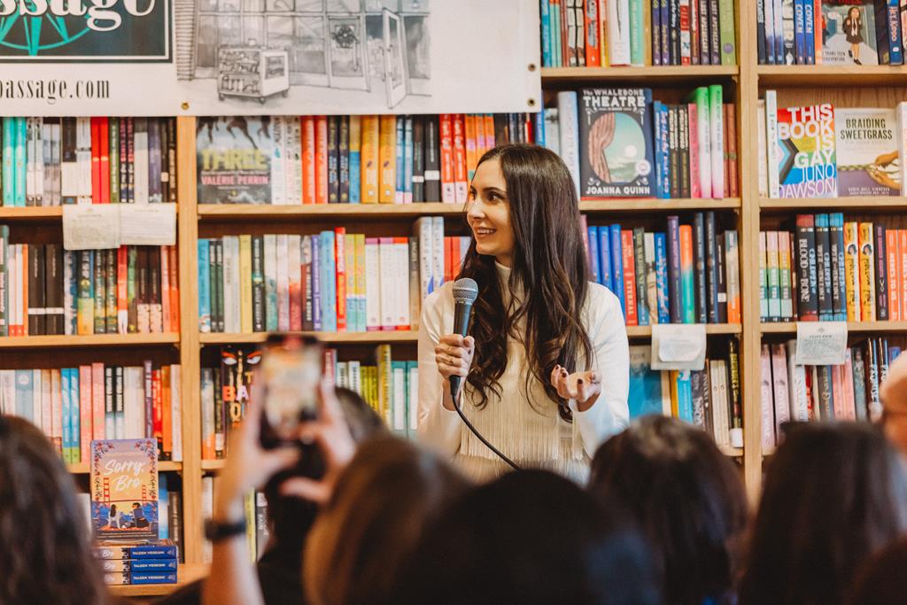 The authour is standing in front of a wall of books and is holding a micrphone, giving a talk to an audience. 