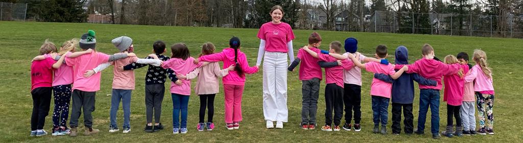 A teacher looking at the camera with 8 children on both sides of her. The teacher and the children are wearing pink.