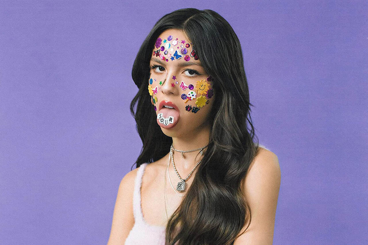 Album art from Olivia Rodrigo's Sour, which shows a woman with children's stickers of butterflies, sun, and similar covering her face. She's sticking out her tongue, and has stickers spelling the word 
