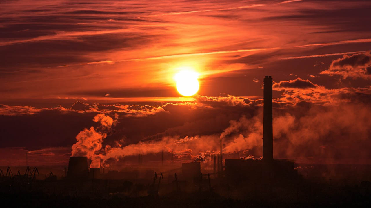 Photo of a red sunset behind a building with many industrial chimneys spitting out thick clouds of smoke.