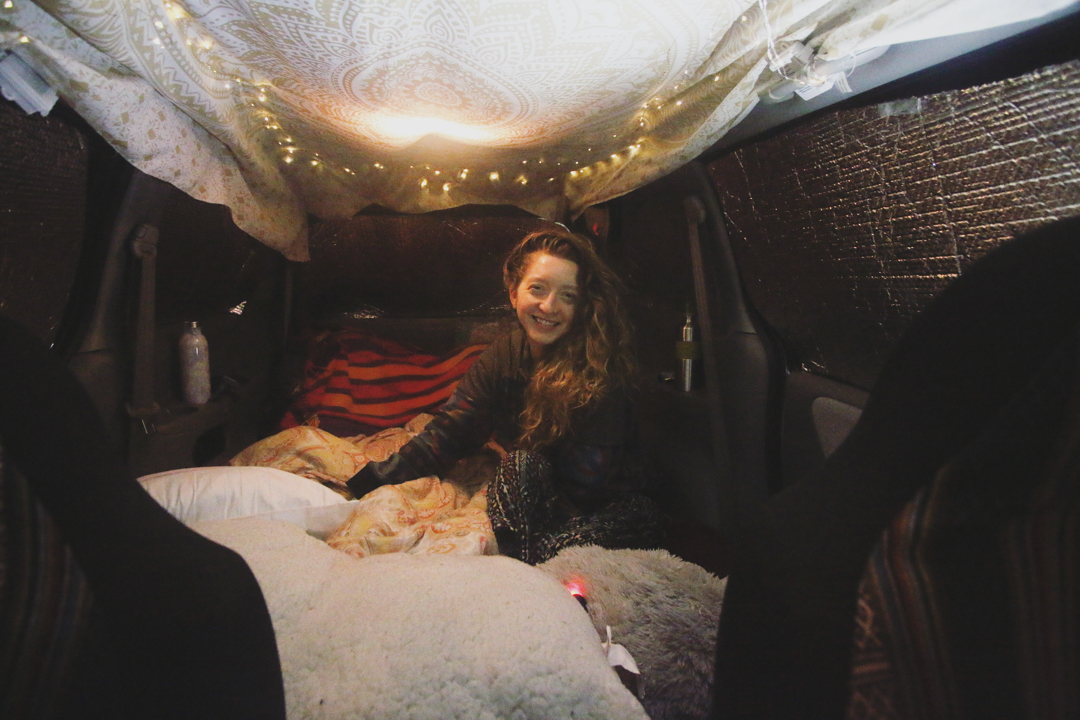 Photo of Andrea Sadowski happily showing the inside of her van home, with filled with blankets and pillows, with fairy lights along the ceiling
