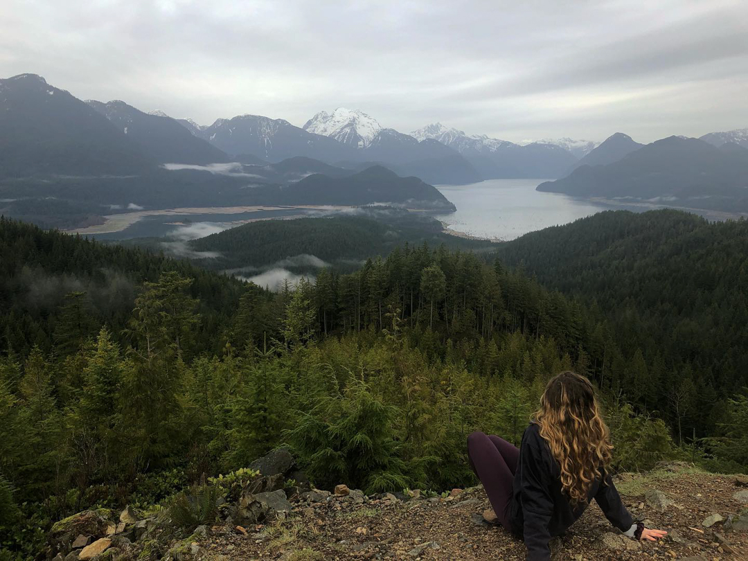 Photo of a person sitting on the edge of a cliff, looking out over forests, lakes, and mountains