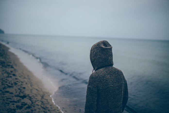 Moody photo of a person walking along a cloudy grey beach with a hoodie pulled up over their head