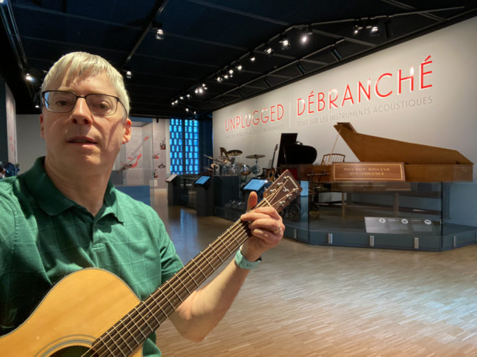Photo of Eric Spalding holding a guitar, standing in front of a display of insturments in a museum