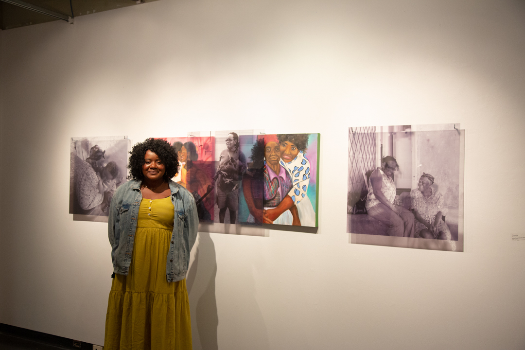 Photo of Natoya Ellis standing in front of a wall showing off their art