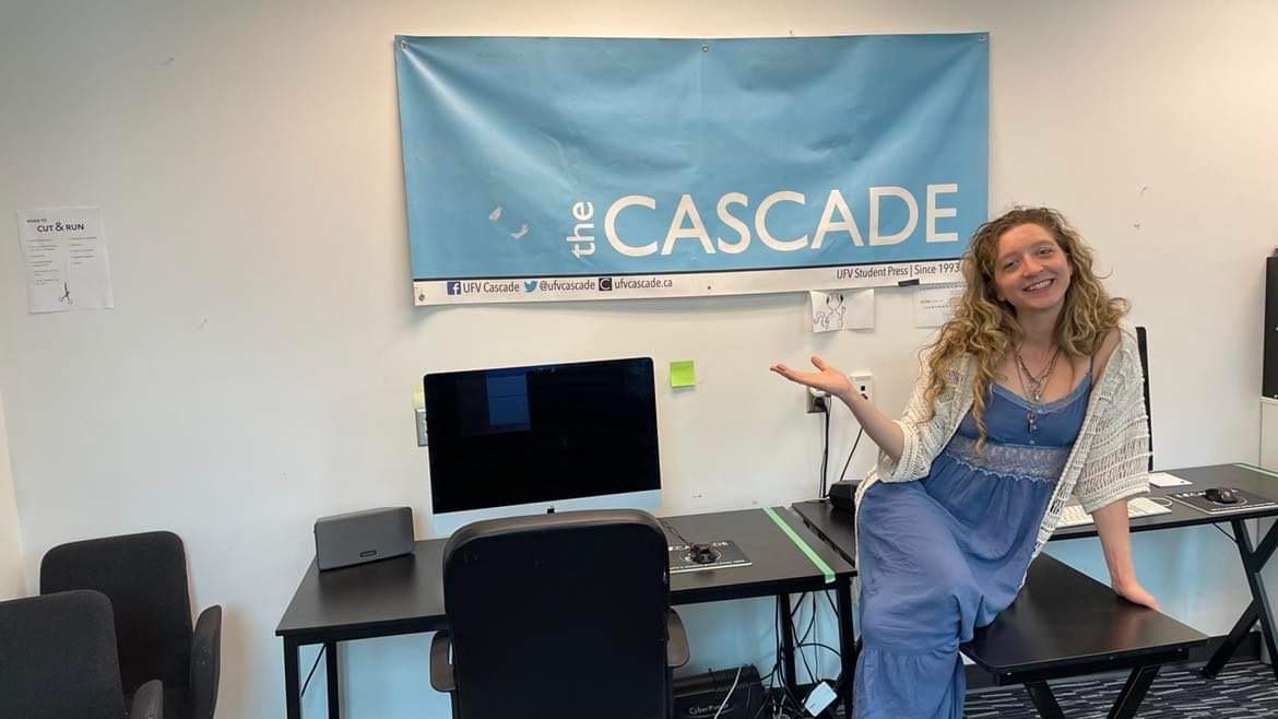 Photo of Andrea Sadowski sitting on a desk in The Cascade's office, gesturing at a banner with our logo on it