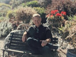 Carl Peters sitting on a bench in nature. Surrounded by red flowers in the sunshine.