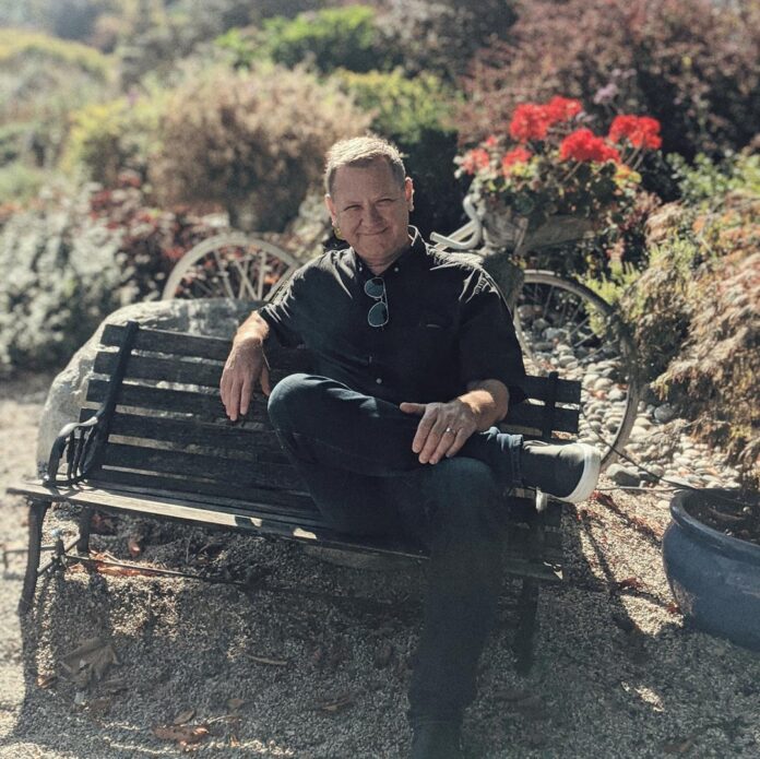 Carl Peters sitting on a bench in nature. Surrounded by red flowers in the sunshine.