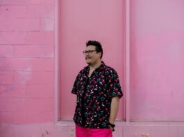 Jordan Abel posing infront of a pink wall wearing a flowery shirt, smiling and looking away from the camera.