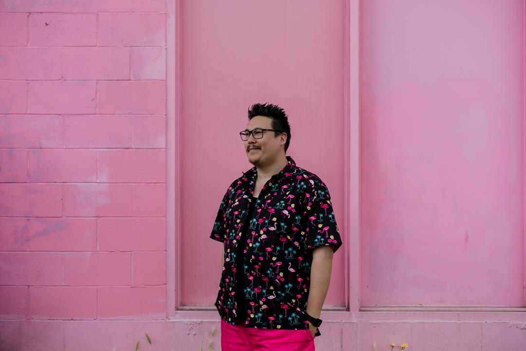 Jordan Abel posing infront of a pink wall wearing a flowery shirt, smiling and looking away from the camera.