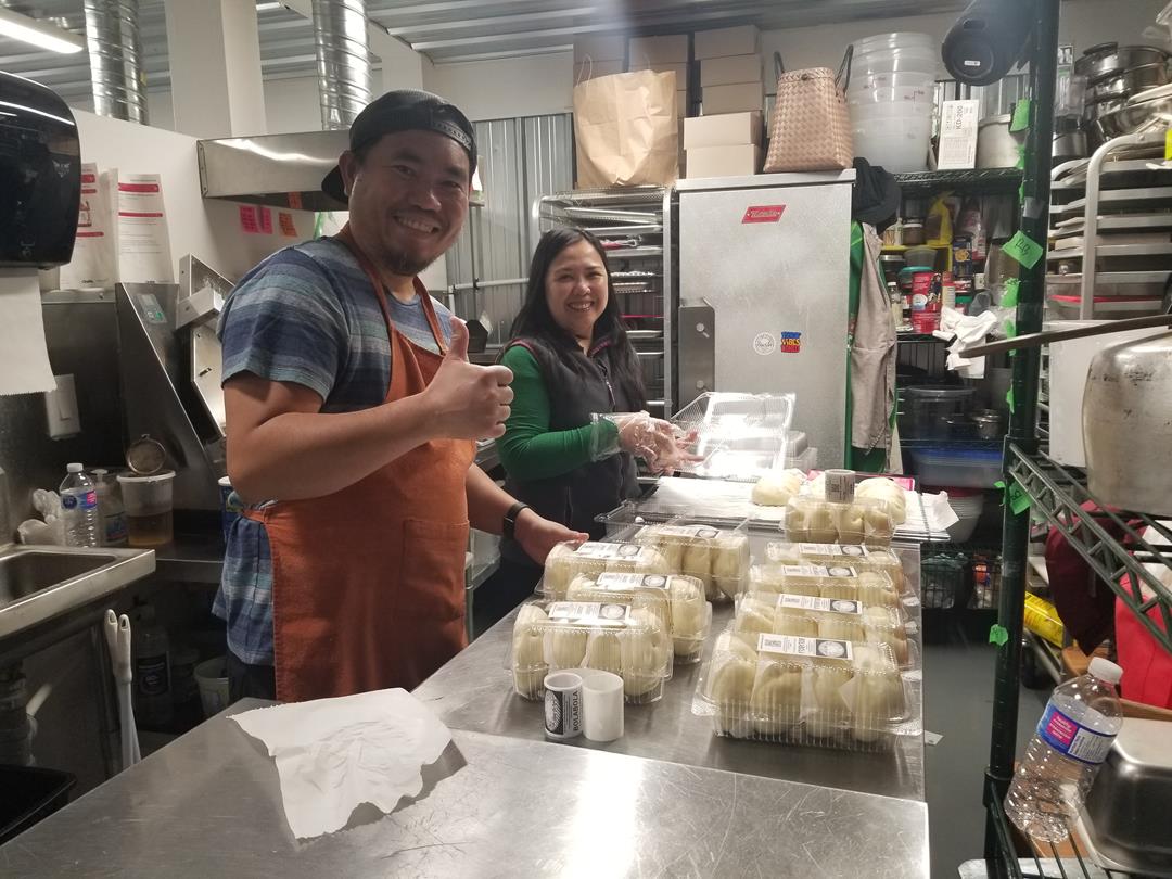 A man and a women in an industrial kitchen making dumplings. They are looking at the camera, smiling and giving thumbs up.