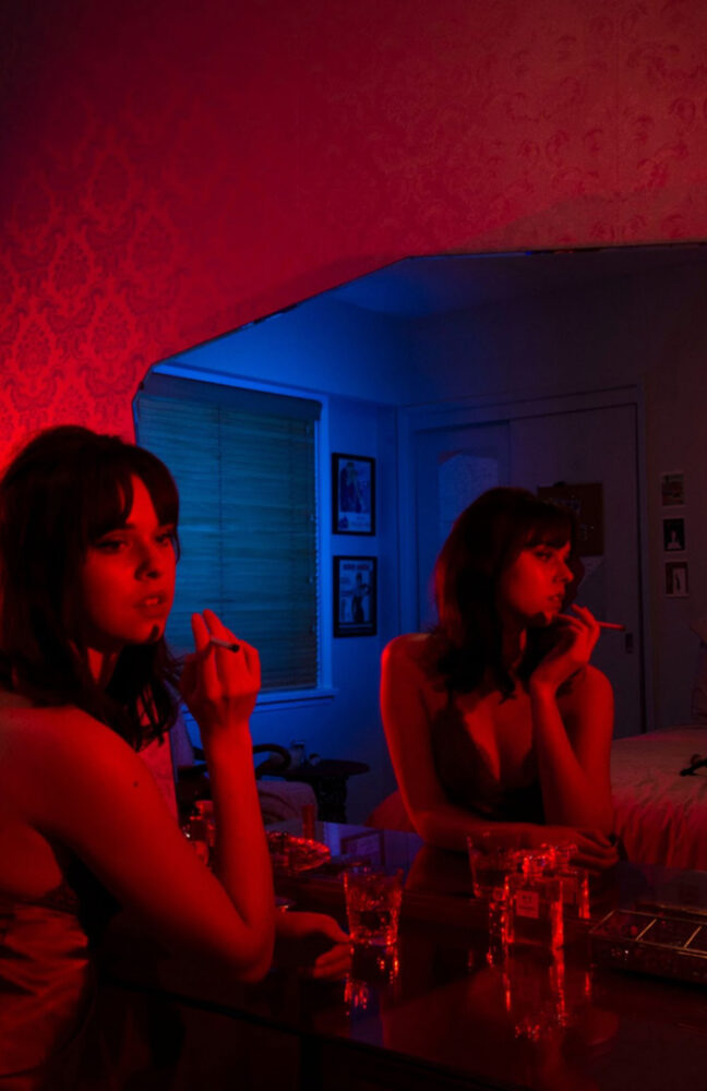 Photo of a person in a nightgown smoking in front of a mirror. They are cast in red light, as is the wall behind the mirror. In the mirror, we see the room behind is a cold blue. On the vanity with the mirror is a glass and a small liquor bottle. The person is looking away from the mirror at something not visible, seemingly lost in thought.