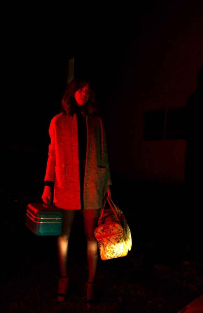 Photo of a person standing in the dark. They are lit by a bright red light from one side, and what looks like car headlights from around knee-level. They are wearing a suit jacket and short skirt, and holding two old-fashioned travel bags as they look towards the light.