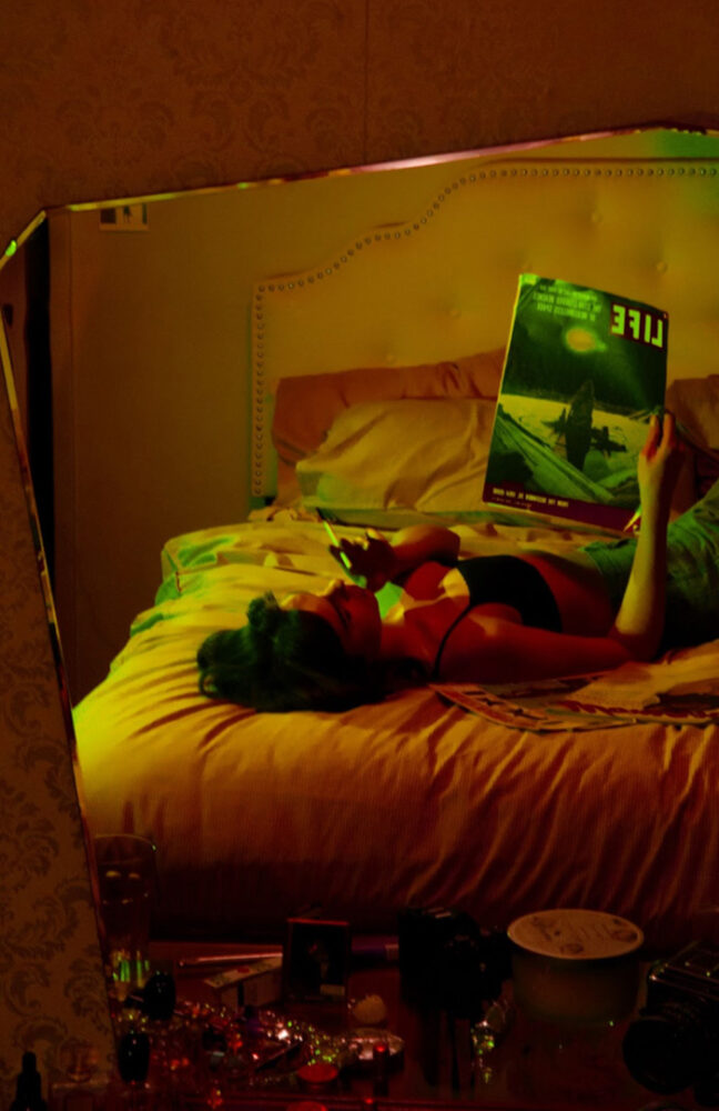Photo of a person reflected in a mirror. There is a strong orange/yellow light throughout the photo. They are lying across the foot of a bed on their back, smoking a cigarette. They are wearing a bra and jeans, and reading "Life" magazine.