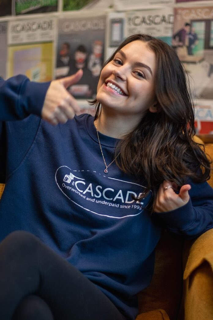 Photo of Sydney Marchand in the Cascade Office. On the wall behind her is previous cascade covers. She sits on a couch and give two thumbs up while smiling. She wears cascade merch reading 