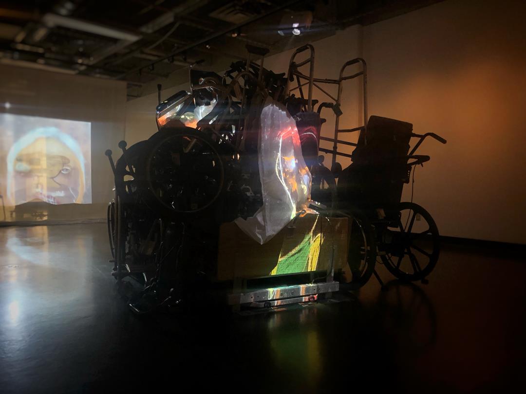 A dark art gallery room, in the center is a pile of equipment like a wheelchair and a walker. There is a projector in the center of the pile and it is projecting images onto the wall across. The images are paintings of abstract faces.