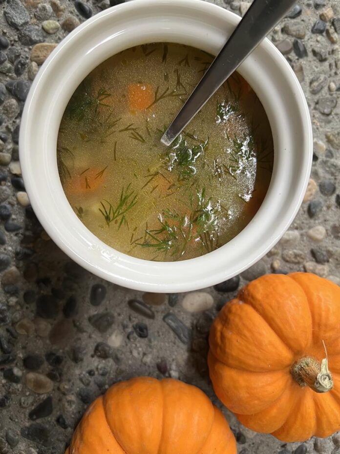 A white bowl filled with orzo soup white a spoon sticking out. Beside the bowl sits some mini orange pumpkins.