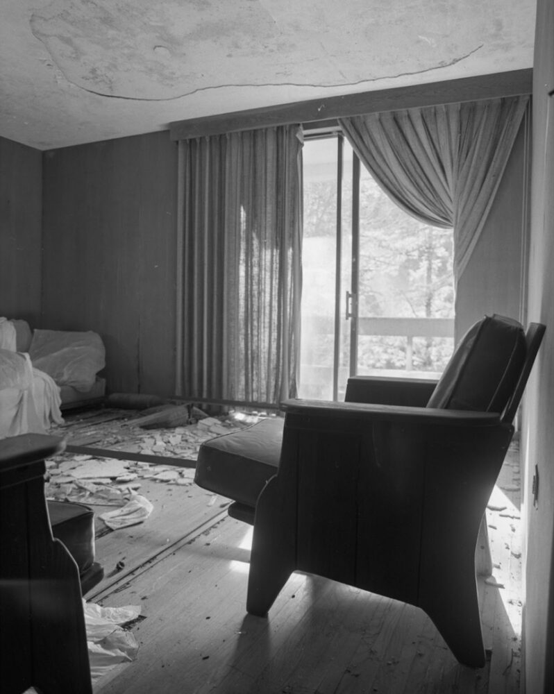 Black and white photo of an abandoned house's interior. The ceiling's plaster seems to be crumbling, and chunks litter the floor. Several pieces of old furniture, including a chair in the foreground, look worn and disused. A balcony door is pulled open, and one of two curtains is pulled outside of the home, letting bright natural light into the dark room.