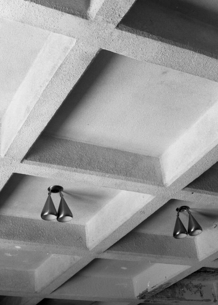 Black and white photo of the ceiling of an abandoned house. Rafters in an unusual pattern segment it into squares. Two of them have small, old-fashioned light fixtures hanging from them.