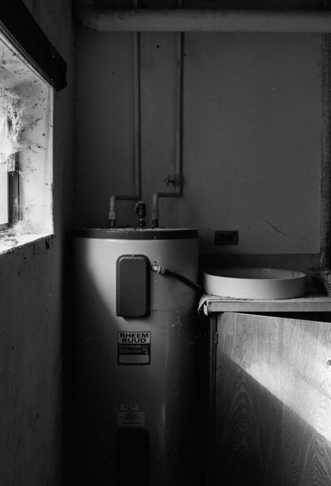 Black and white photo of a hot water tank in an old abandoned house. A small window is filled with cobwebs, and light from it cast harsh shadows on the scene.