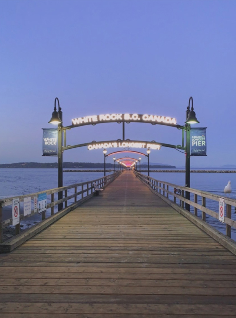 Photo looking directly down a pier just before dawn. A sign above it reads in florescent text: "White Rock B.C. Canada. Canada's longest pier." Two blue banners flanking that sign repeat the message. Further down, more lights illuminate the pier, seeming to get closer and closer in the distance until they become one red and yellow glow.