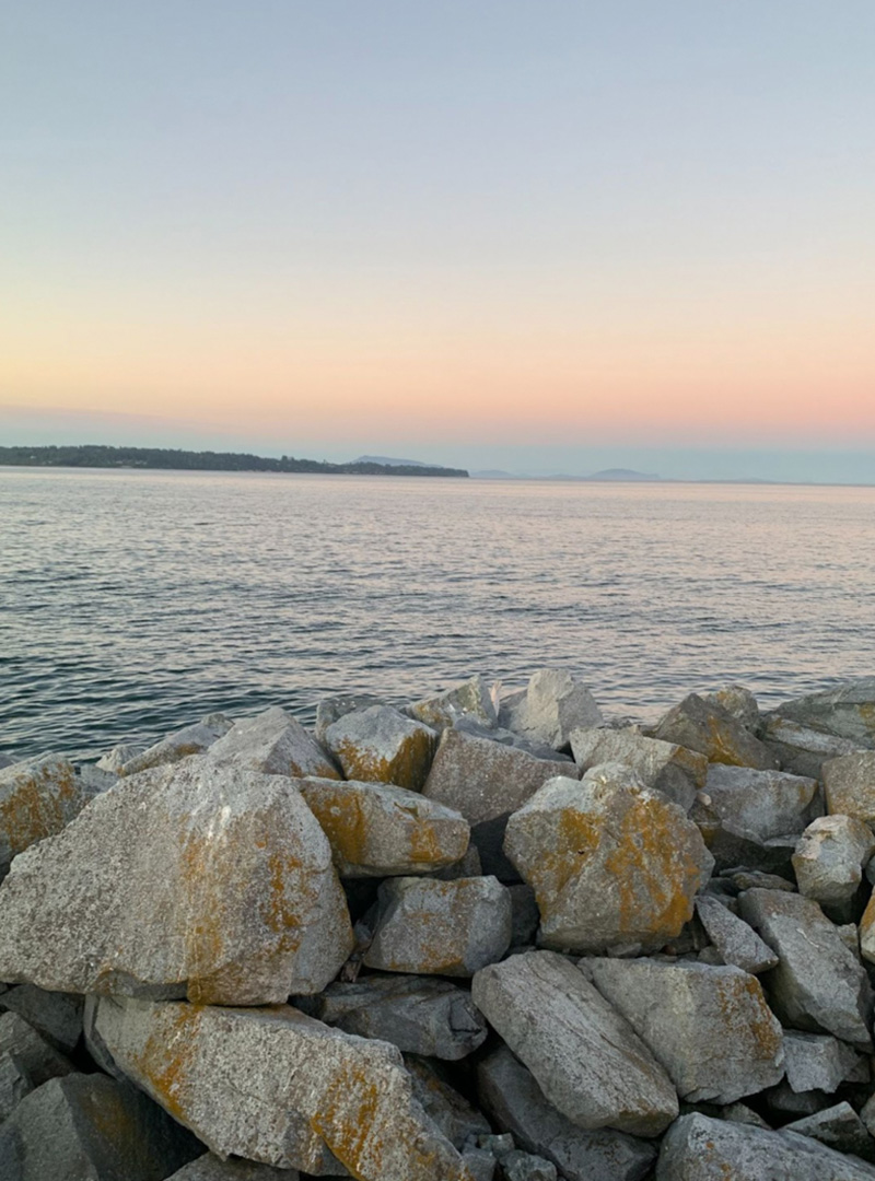 Photo of a pile of large rocks next to the ocean on a beach. The camera is looking out across the ocean, where pinkish clouds make it clear the sun is rising.
