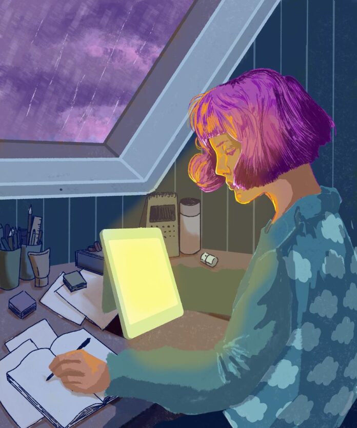 A woman with pink hair sitting a desk in a dark room facing a window showing a rainy scene outside. On her desk sits a SAD lamp radiating a warm light onto her face