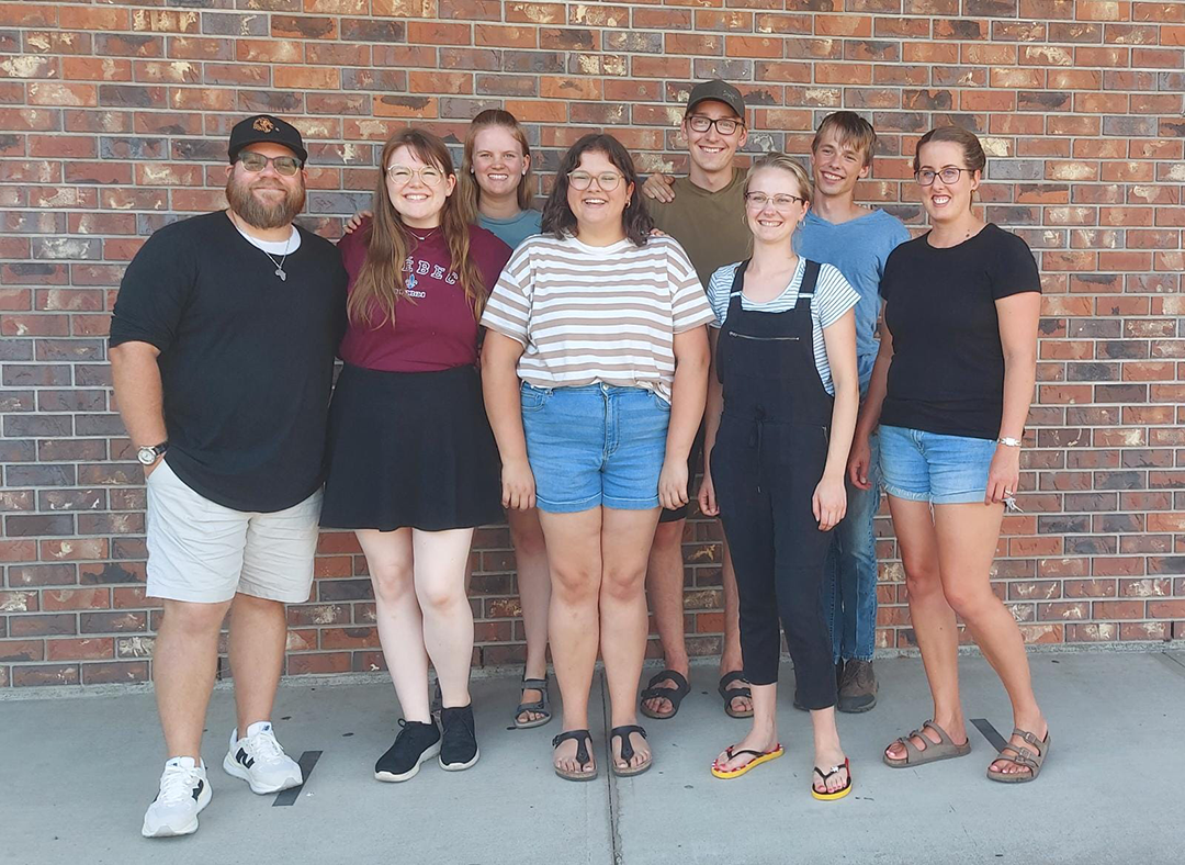 The UCM club group stands in a row in front of a brick wall on a sunny day.