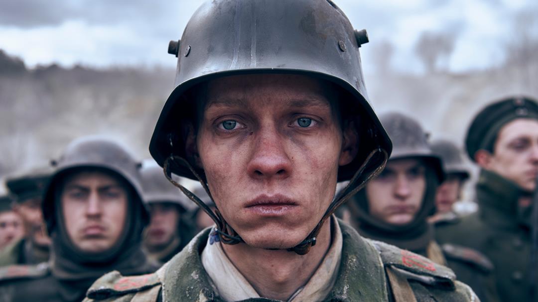 A group of soliders stand outside. There faces are visible. One man stands out while the rest are blurred. He wears an old style army helmet with dirt on his face. He is causacian with blue eyes and looks weary.