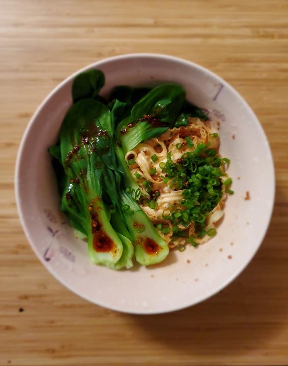 A bowl of noodles topped with baby bok choy and scallions drizzeld with chili oil.