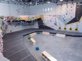 Photo of the rock climbing gym with rock walls surrounding the space. The rocks are different bright colors and in the center on the ground are black soft mats.
