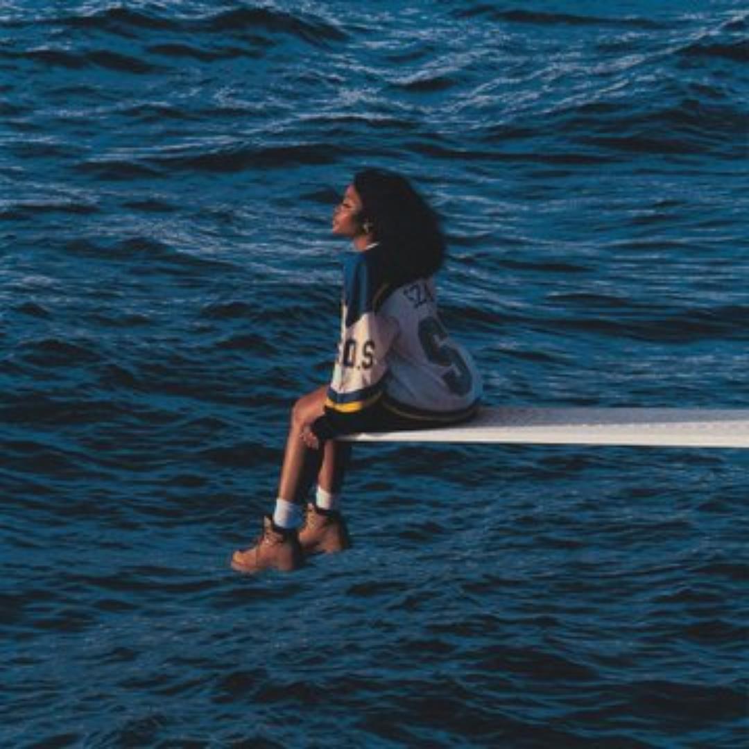 SZA sitting on a diving board over the ocean, all that can be seen is the board, seemingly floating over the blue ocean. She is wearing a sports jersey