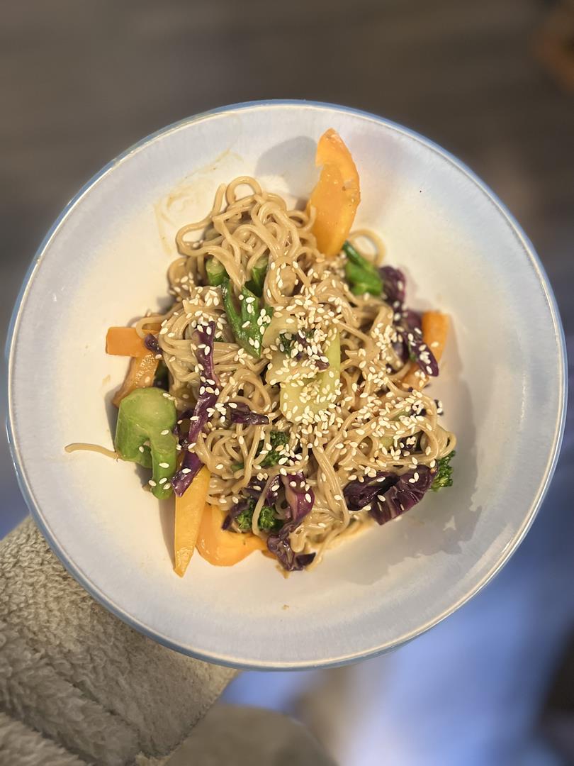 Photo of white bowl being held. Inside is the noodle dish with purple cabbage, broccoli, yellow peppers and noodles topped with sesame seeds.