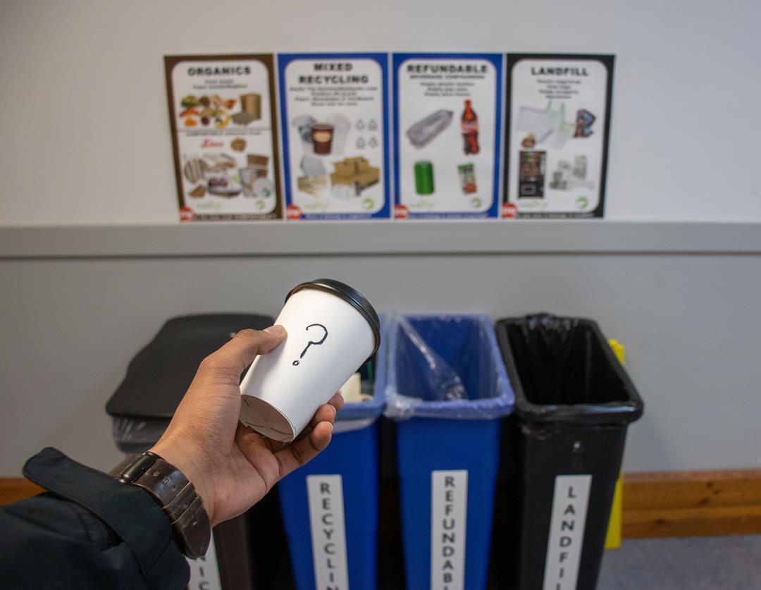 A photo of a hand holding a disposable coffee cup with a question mark drawn on it in front of the 4 types of garbage bins at UFV.