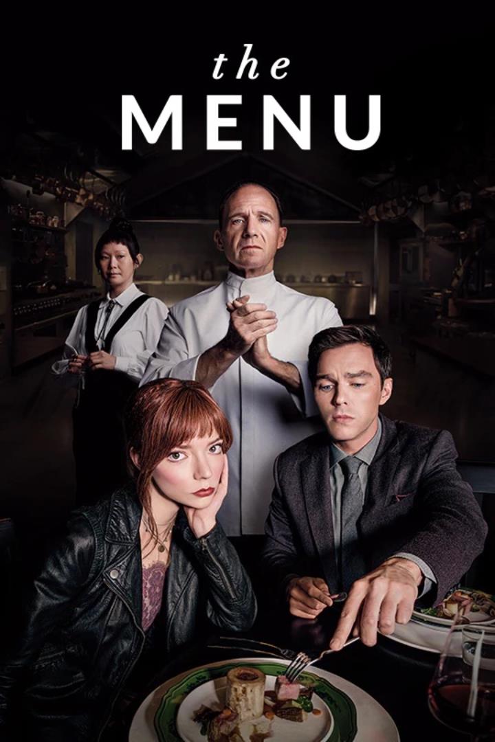 A man and a woman sit at a table the man is reaching for a bite of a dinner plate while the girl stares at the camera. Behind them is a chef, and behind them is a waiter. In the background is a dark professional kitchen.