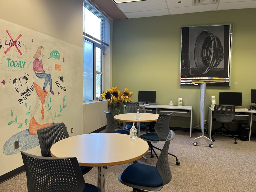 A photo of the academic success centre at UFV in room G126. There are some tables and chairs with computer desks along the back wall. To the left is a large white board with drawings all over it.