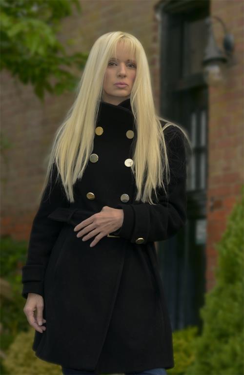 A photo of Jay Lang standing in front of a brick wall. She has long blonde hair and it wearing a double breasted black coat
