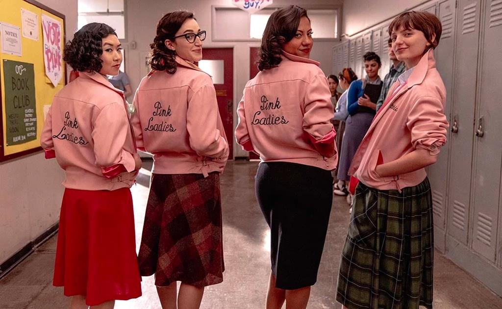 The four pink ladies stand in a line in the hallway with their backs turned to the cameras their heads are all turned looking at the camera and they are wearing pink satin jackets with 