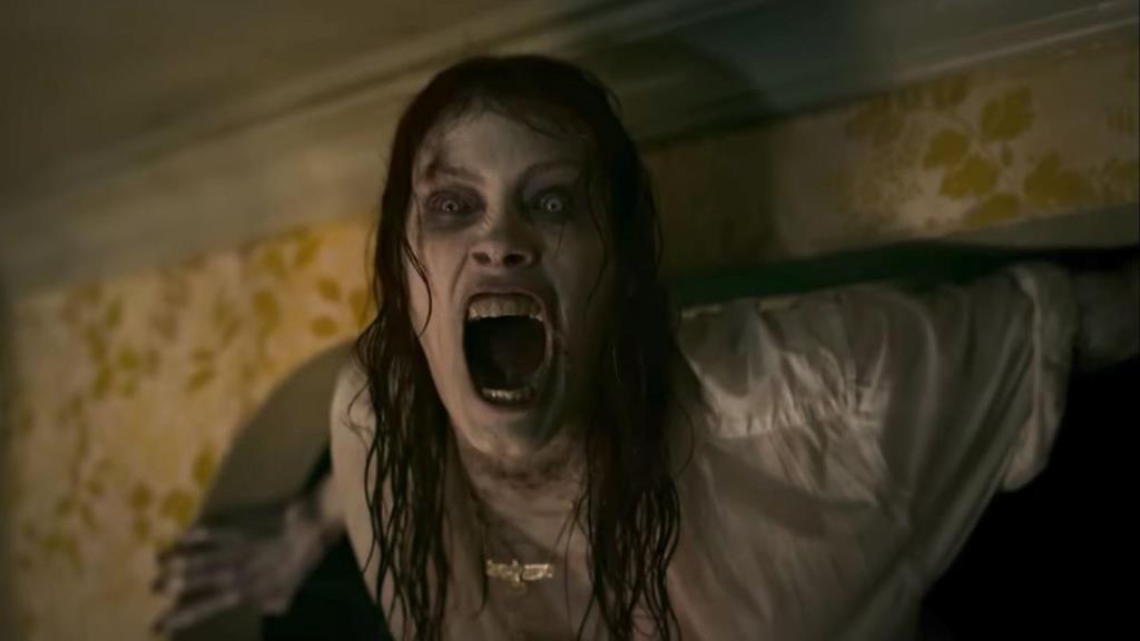 A scene from the movie, an evil woman is standing against the wall in a dark room. She has pale skin and looks to be screaming.
