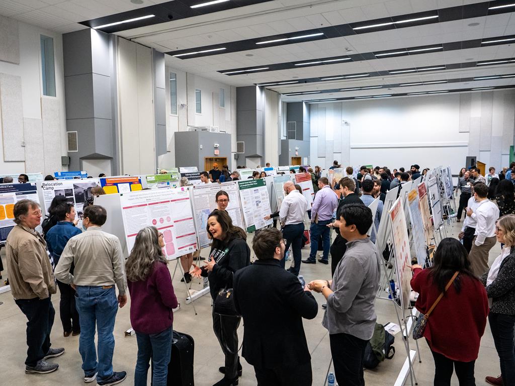 A photo of the research day, in a large room with a crod looking at various posters showcsaing the students research work.