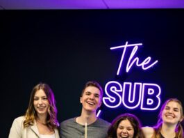 Members of the SUS team, including Ashley McDougall stand behind a pool table in the SUS lounge. There is a neon purple sign behind them that says "The Sub"